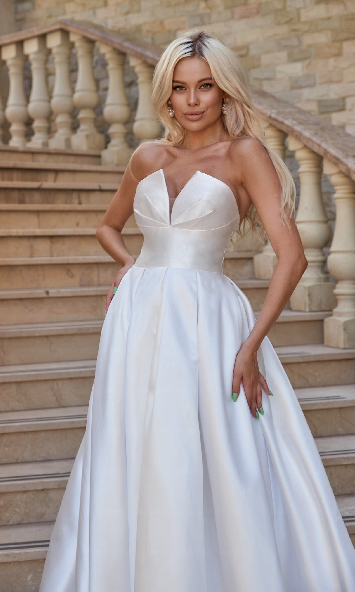 A woman poses gracefully on stone steps with a hand on her hip, wearing the Strapless Off-White Bridal Gown: Tina Holly TA611W in front of a balustrade.
