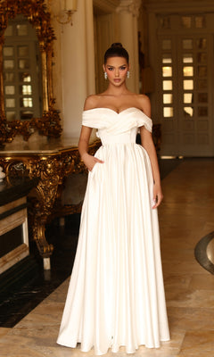 A woman stands indoors near ornate gold decor and large mirrors, wearing the Off-White Long Prom Ball Gown: Tina Holly TE207W—an elegant, floor-length off-the-shoulder dress.