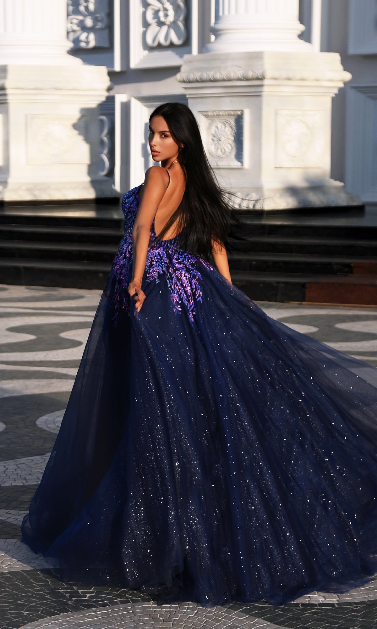 A woman with long dark hair wears the Tina Holly TE216B long formal dress, a deep blue glitter tulle gown with purple floral appliques, standing outdoors before ornate white columns.