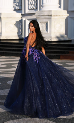A woman with long dark hair wears the Tina Holly TE216B long formal dress, a deep blue glitter tulle gown with purple floral appliques, standing outdoors before ornate white columns.