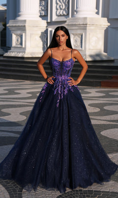 A woman stands outside a white classical building wearing the Tina Holly TE216B, a dark blue glitter tulle ball gown with purple floral appliques and thin straps.