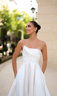 A woman wears the Strapless Long White Prom Dress: Tina Holly TE302 outdoors, smiling and looking to the side with trees and a building in the background.