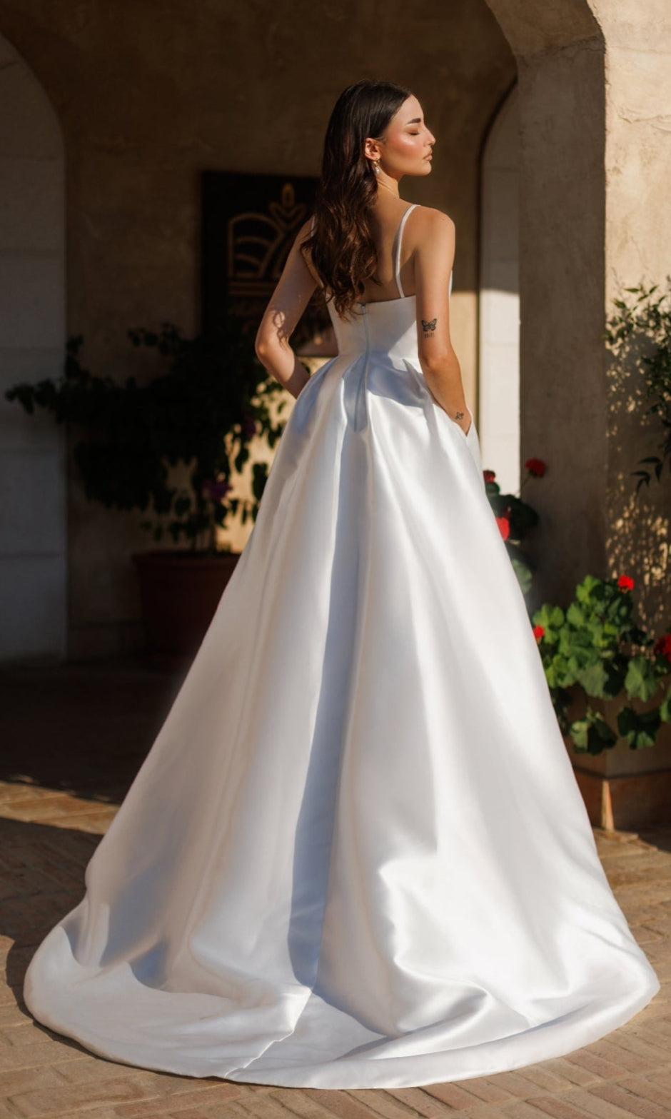 A woman in the Classic Long White Formal Dress: Tina Holly TE305 stands outdoors under an archway with plants nearby, hands in her dress pockets, facing away.