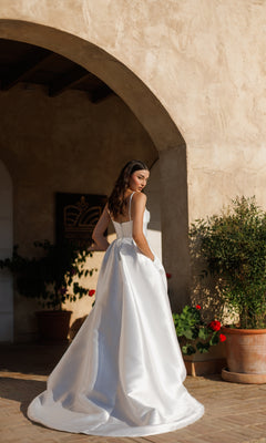A woman in the Classic Long White Formal Dress: Tina Holly TE305 stands outdoors by an arched stone wall, looking over her shoulder.