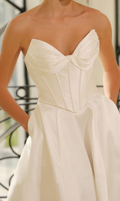 Woman wearing the Strapless Mikado Wedding Dress: Tina Holly TE704, featuring a structured, pleated bodice and full skirt, stands near a decorative black railing.