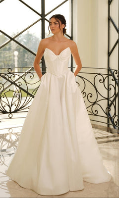 A woman in the Tina Holly TE704 strapless mikado wedding dress in off white, featuring a fitted bodice and full skirt, stands indoors by a decorative metal railing and large windows.