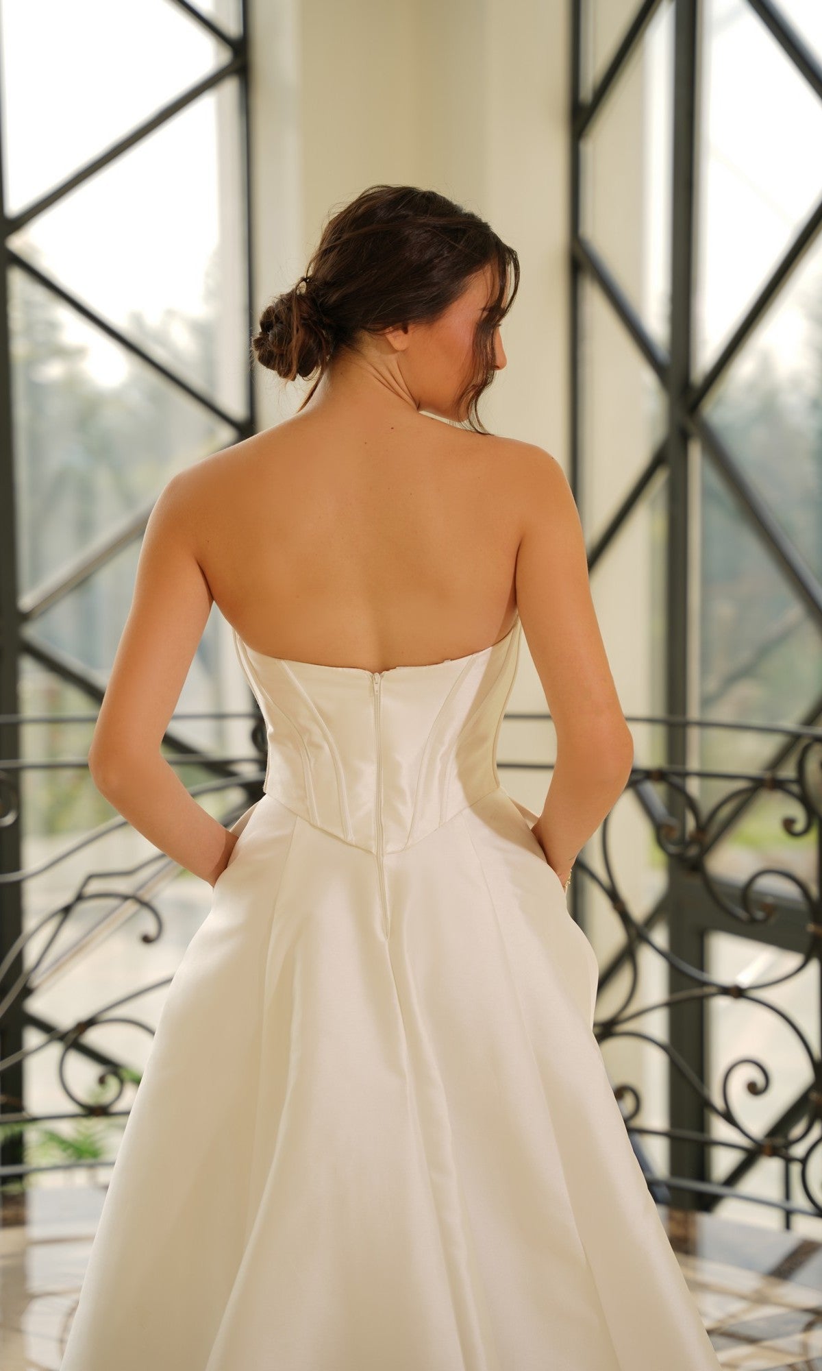 A woman stands indoors with her back to the camera, hands in pockets, wearing the Strapless Mikado Wedding Dress: Tina Holly TE704, framed by large windows featuring geometric patterns.