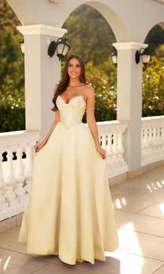 A woman stands outdoors before white columns, wearing the Tina Holly TE704P long formal dress in light yellow, featuring a strapless fitted bodice and a full skirt.