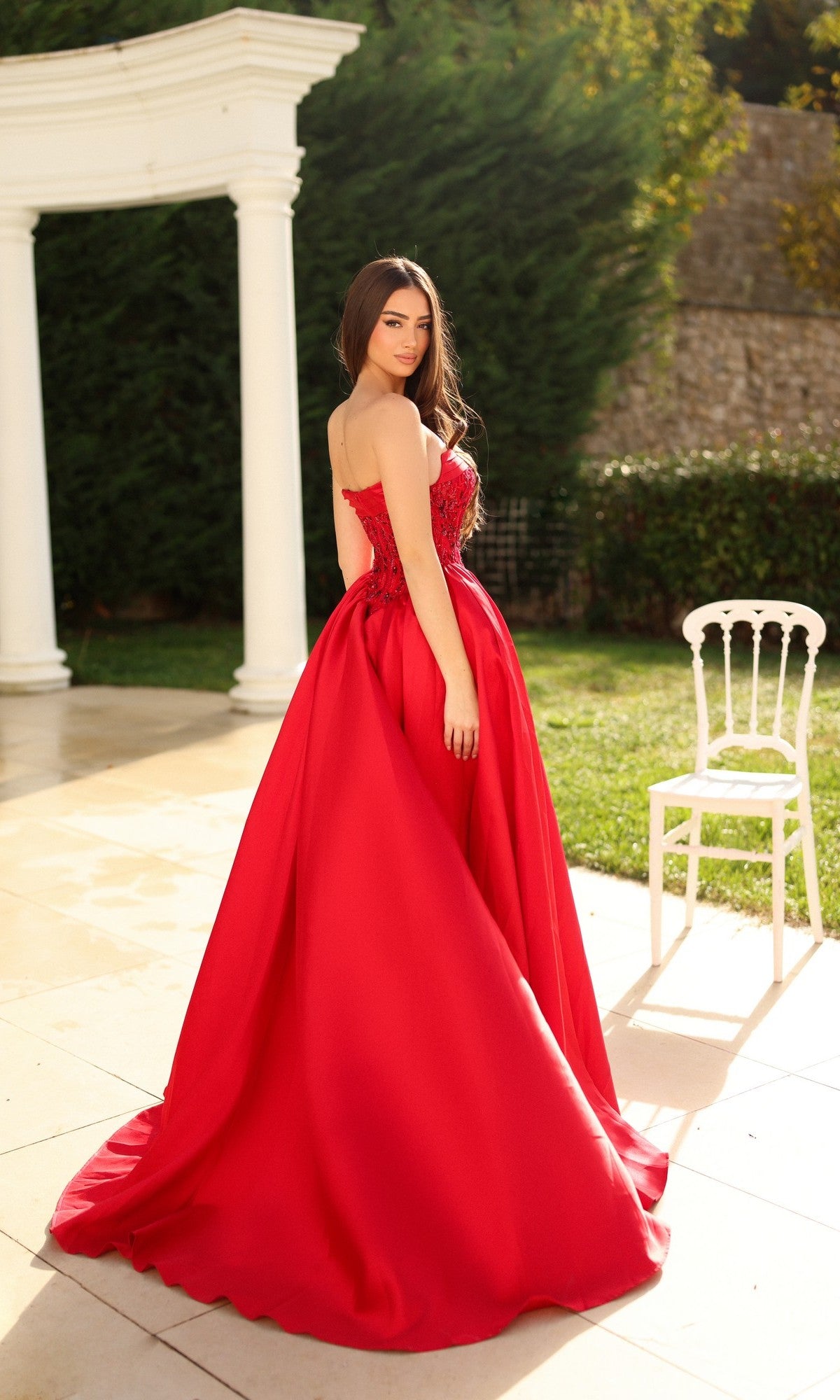 A woman wearing the Long Formal Dress: Tina Holly TE706, featuring a strapless embroidered corset bodice, stands outdoors on a patio near a white chair and classical columns with greenery in the background.