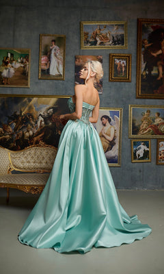 A woman wearing the Tina Holly TE706 long formal dress in light blue stands before a wall of classical paintings and a vintage sofa, glancing over her shoulder.