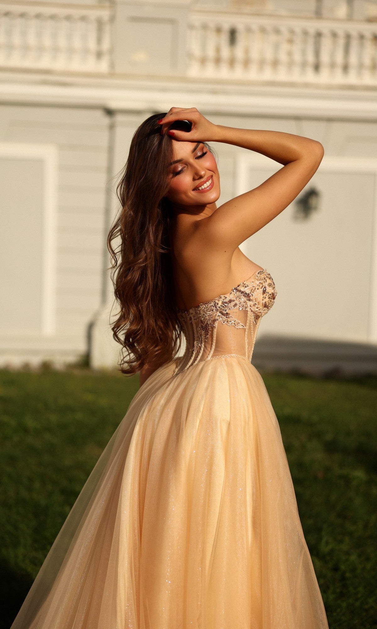 A woman smiles outdoors on grass in the Long Formal Dress: Tina Holly TE708, a strapless glitter ball gown, with one hand touching her hair.