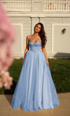 A woman in the Long Formal Dress: Tina Holly TE708, a strapless glitter ball gown with a sheer corset bodice, stands outdoors on a paved area with green grass and an ornate building enhancing the elegant scene.