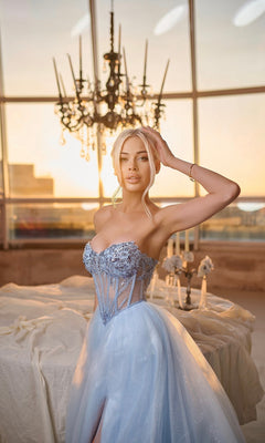 A woman in the Tina Holly TE708 long formal dress—a pale blue strapless glitter ball gown with a lace corset bodice—stands indoors by a table, under a sparkling chandelier and sunset-lit windows.