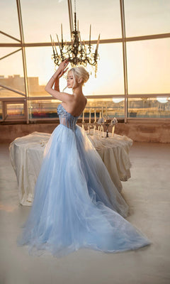 A woman wearing the Long Formal Dress: Tina Holly TE708— a strapless glitter ball gown with a corset bodice— stands indoors by a white-covered table, adjusting her hair as large windows and a chandelier appear in the background.