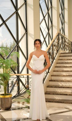 A woman wearing the Contemporary Wedding Dress: Tina Holly TE709 stands at the base of a staircase with large windows and a decorative railing in the background.