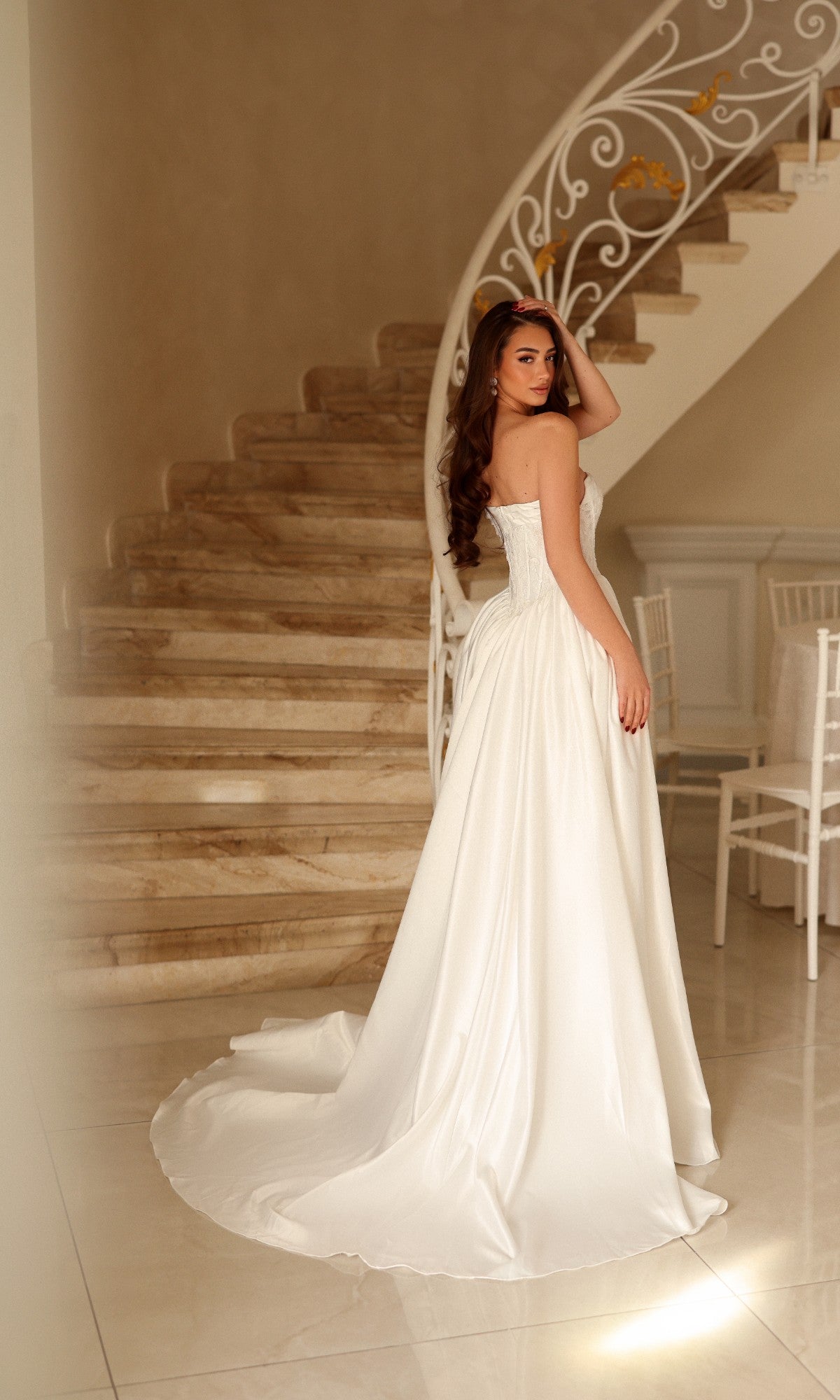 A woman wearing a Tina Holly TE711 strapless, floor-length white wedding dress with a full skirt and train, standing in front of a curved staircase.