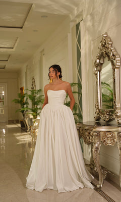 A woman in the Long Off-White Satin Dress: Tina Holly TE810 stands in an elegant hallway with ornate decor, a large mirror, and potted plants.