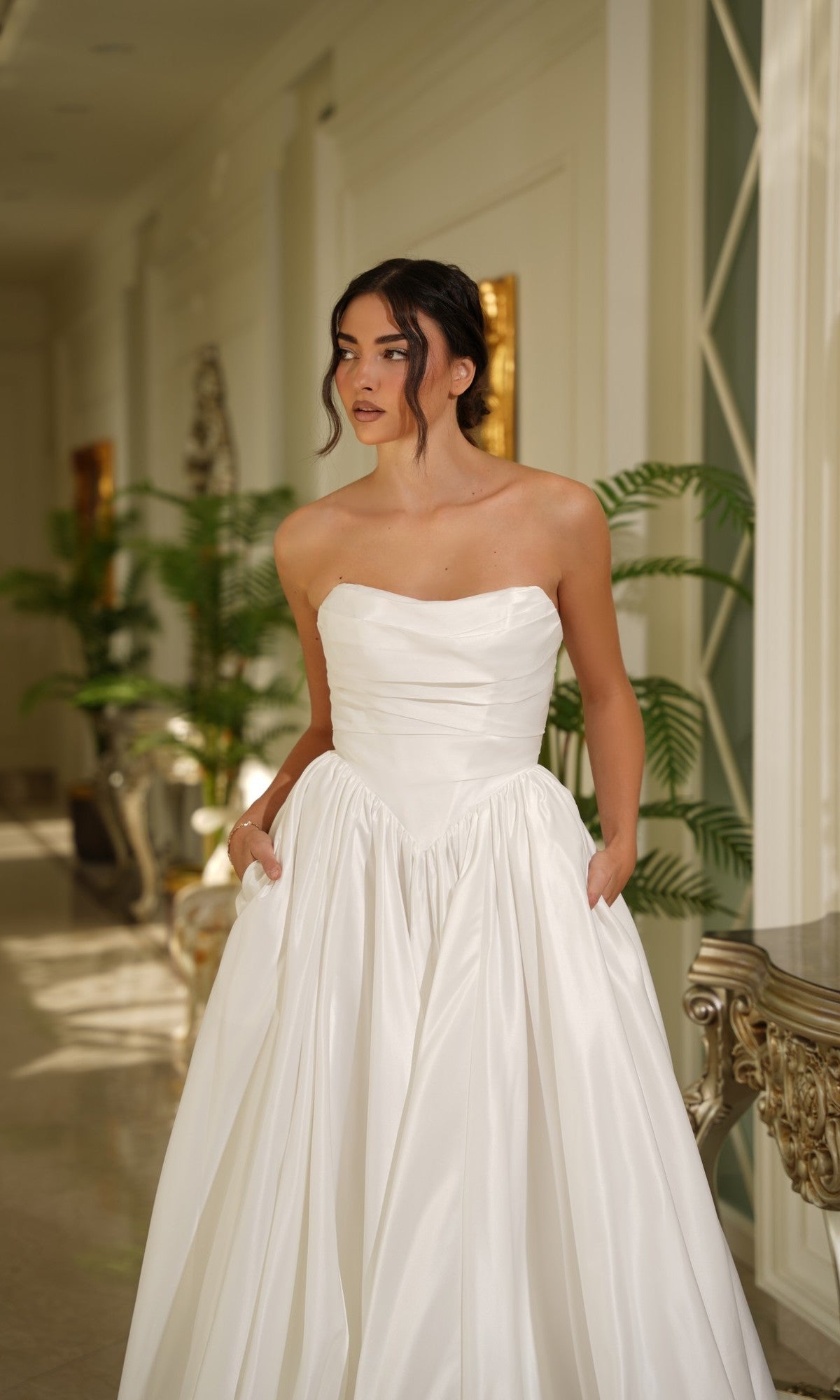 A woman stands indoors in the Long Off-White Satin Dress: Tina Holly TE810, featuring a strapless design and basque waistline with pockets. Green plants and ornate furniture decorate the background.