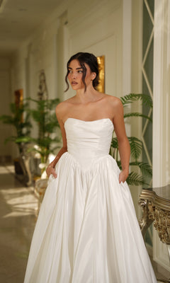 A woman stands indoors in the Long Off-White Satin Dress: Tina Holly TE810, featuring a strapless design and basque waistline with pockets. Green plants and ornate furniture decorate the background.