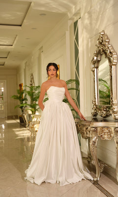 A woman wearing the Long Off-White Satin Dress: Tina Holly TE810 stands in an elegant hallway with marble floors, ornate furniture, and greenery.