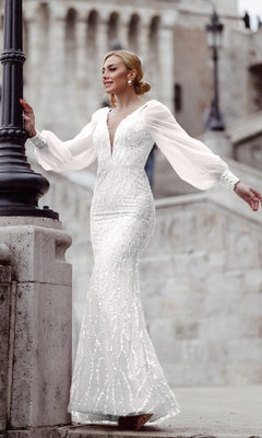 A woman in the Tina Holly TK209 Long Sleeve Long Prom Dress, adorned with beaded details, stands by a stone railing outdoors, the embellished gown shimmering against the blurred backdrop of a historical building.