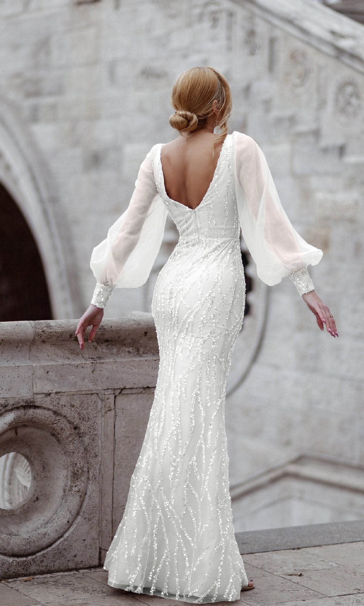 A woman in the Tina Holly TK209 long sleeve long prom dress stands on a stone staircase, facing away with her hair in a low bun.