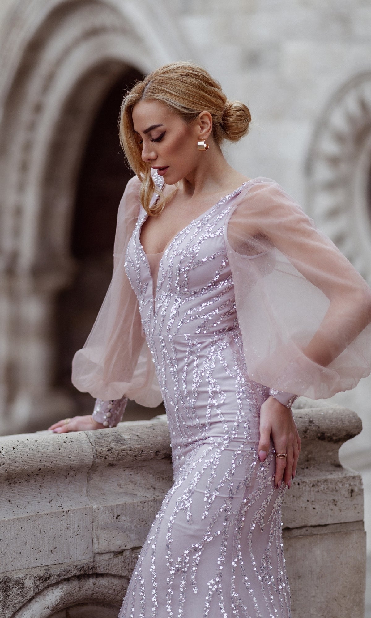 A woman wearing the Tina Holly TK209 Long Sleeve Long Prom Dress stands by a stone railing, looking down, with arched stone architecture in the background.