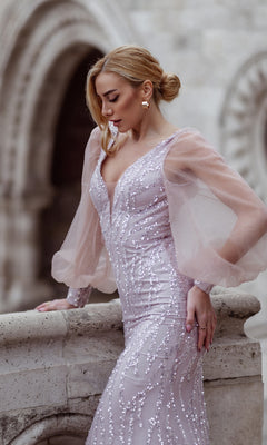 A woman wearing the Tina Holly TK209 Long Sleeve Long Prom Dress stands by a stone railing, looking down, with arched stone architecture in the background.