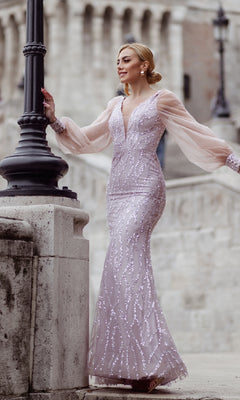 A woman stands outdoors near a stone railing and lamp post wearing the Tina Holly TK209, a long sleeve, form-fitting light pink embellished prom dress.