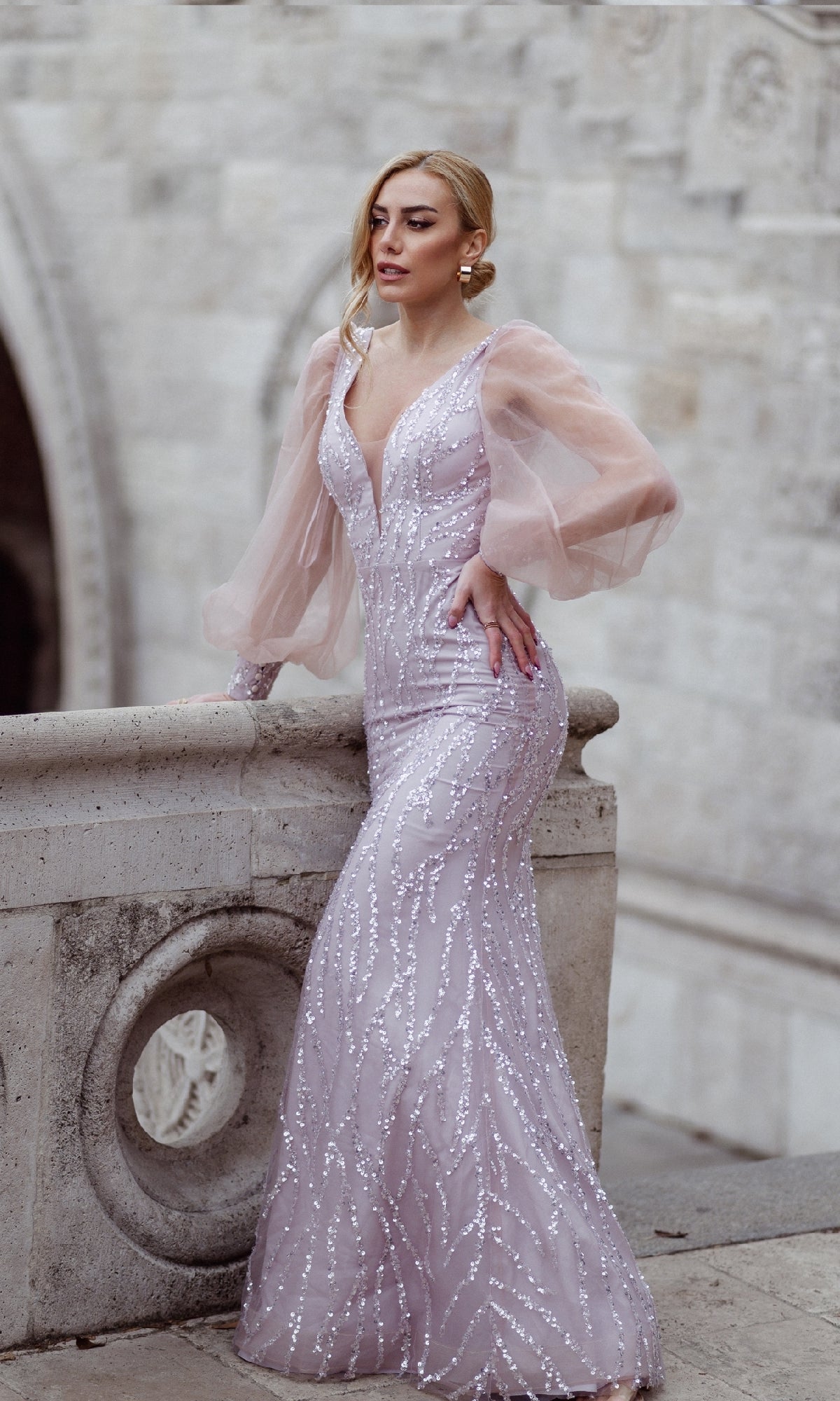 A woman in the Tina Holly TK209 Long Sleeve Long Prom Dress, a fitted light purple gown with sheer puffed sleeves and embellishments, stands by a stone railing with a textured stone wall behind her.
