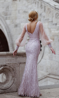 A woman in the Tina Holly TK209 Long Sleeve Long Prom Dress, a floor-length embellished gown in light purple with sheer sleeves, stands on stone stairs facing away from the camera.
