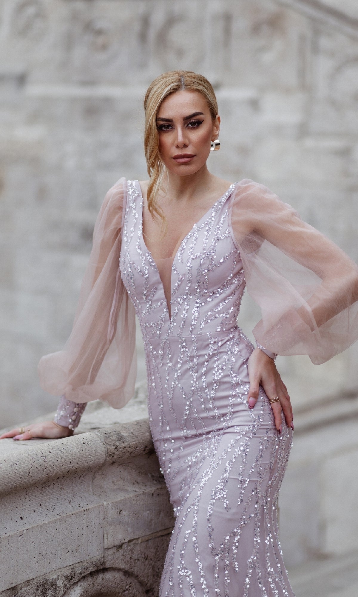 A woman wearing the Tina Holly TK209 Long Sleeve Long Prom Dress stands by a stone railing, looking directly at the camera.