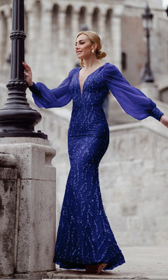 A woman wearing the Long Sleeve Long Prom Dress: Tina Holly TK209, fitted and deep blue with beaded details, stands outdoors by a stone railing and lamppost.