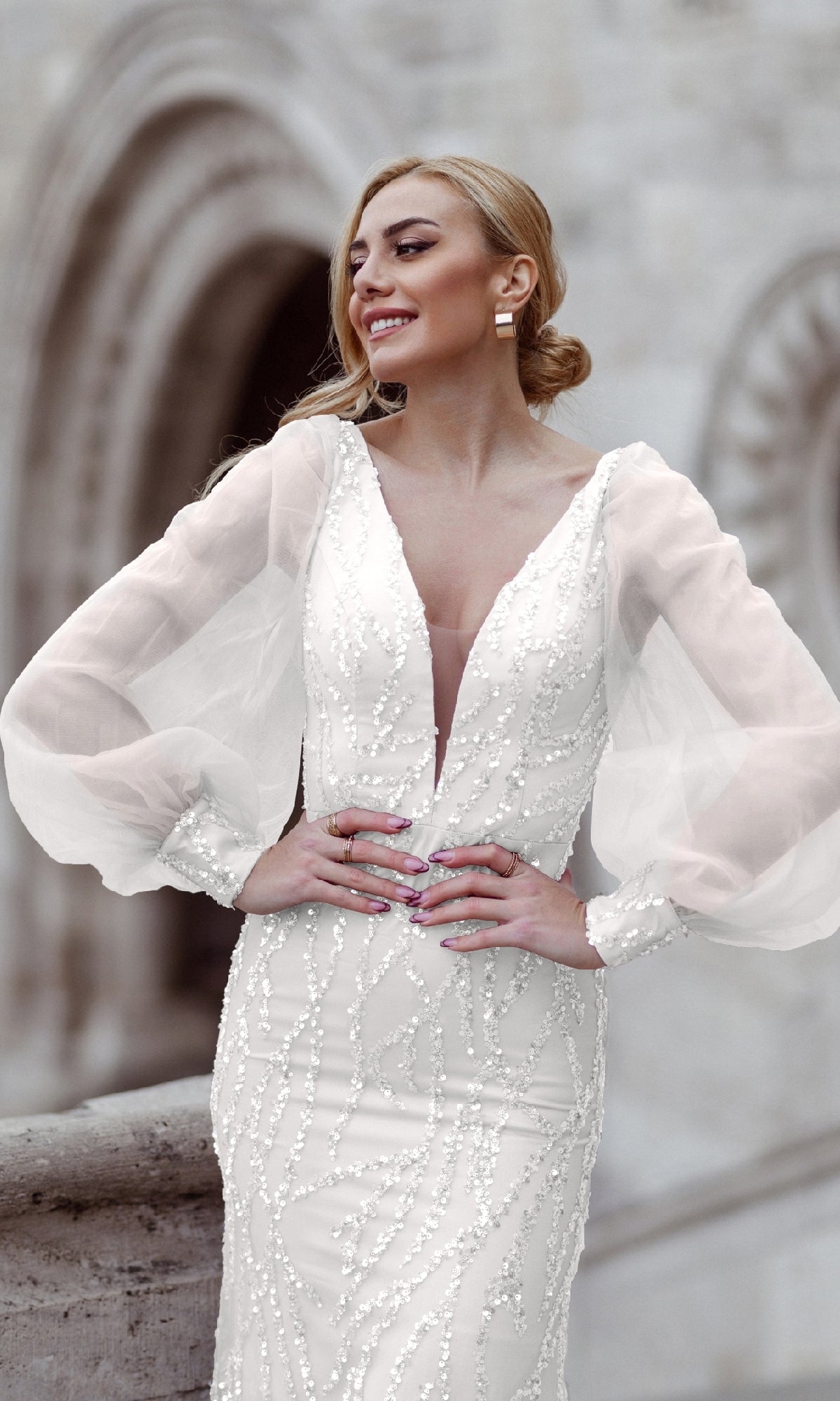 A woman outdoors smiles and looks to the side while wearing the Tina Holly TK209 Long Sleeve Long Prom Dress in white with beaded details.