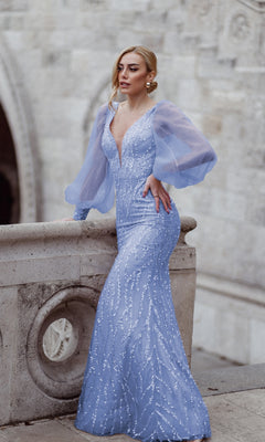 A woman wearing the Tina Holly TK209 Long Sleeve Long Prom Dress, a fitted light blue embellished gown with sheer sleeves, stands by a stone railing against an ornate stone wall.