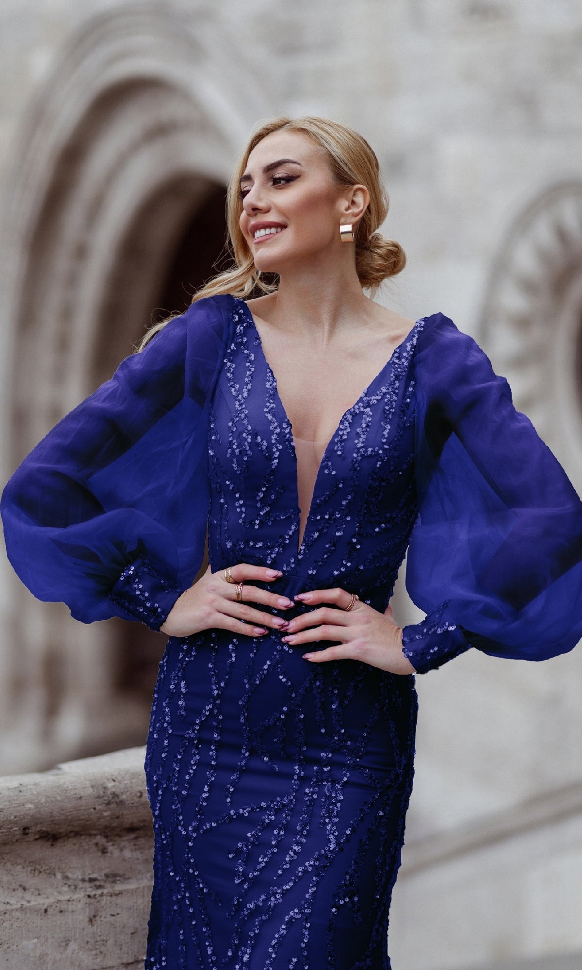 A woman stands outdoors, smiling in the Tina Holly TK209 Long Sleeve Long Prom Dress, featuring a deep blue hue and sheer puff sleeves, with a blurred stone archway in the background.