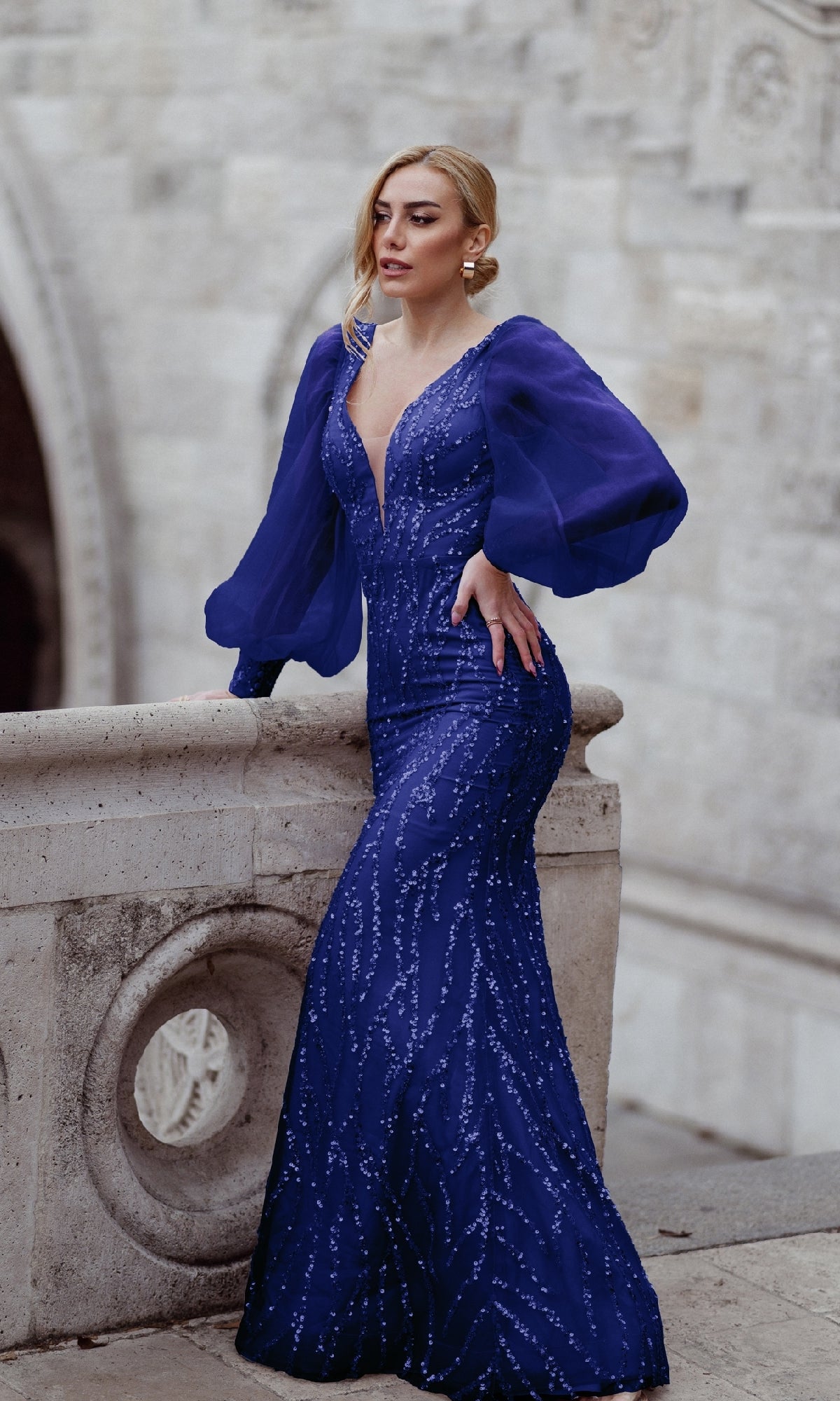 A woman in the Tina Holly TK209 long sleeve prom dress, featuring a fitted deep blue embellished design and sheer puffed sleeves, stands by a stone railing against a textured light wall.