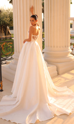 A woman in the Strapless Off-White Formal Dress: Tina Holly TK241, featuring a long train, stands before large white columns and looks over her shoulder.