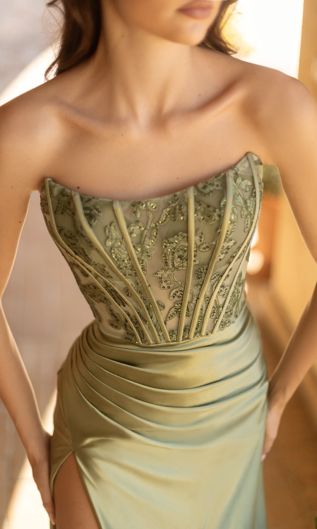 A woman stands indoors wearing the Strapless Long Satin Prom Dress: Tina Holly TK551, featuring a light green color, embroidered bodice, and thigh-high slit.