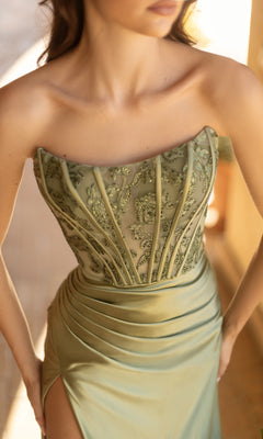 A woman stands indoors wearing the Strapless Long Satin Prom Dress: Tina Holly TK551, featuring a light green color, embroidered bodice, and thigh-high slit.