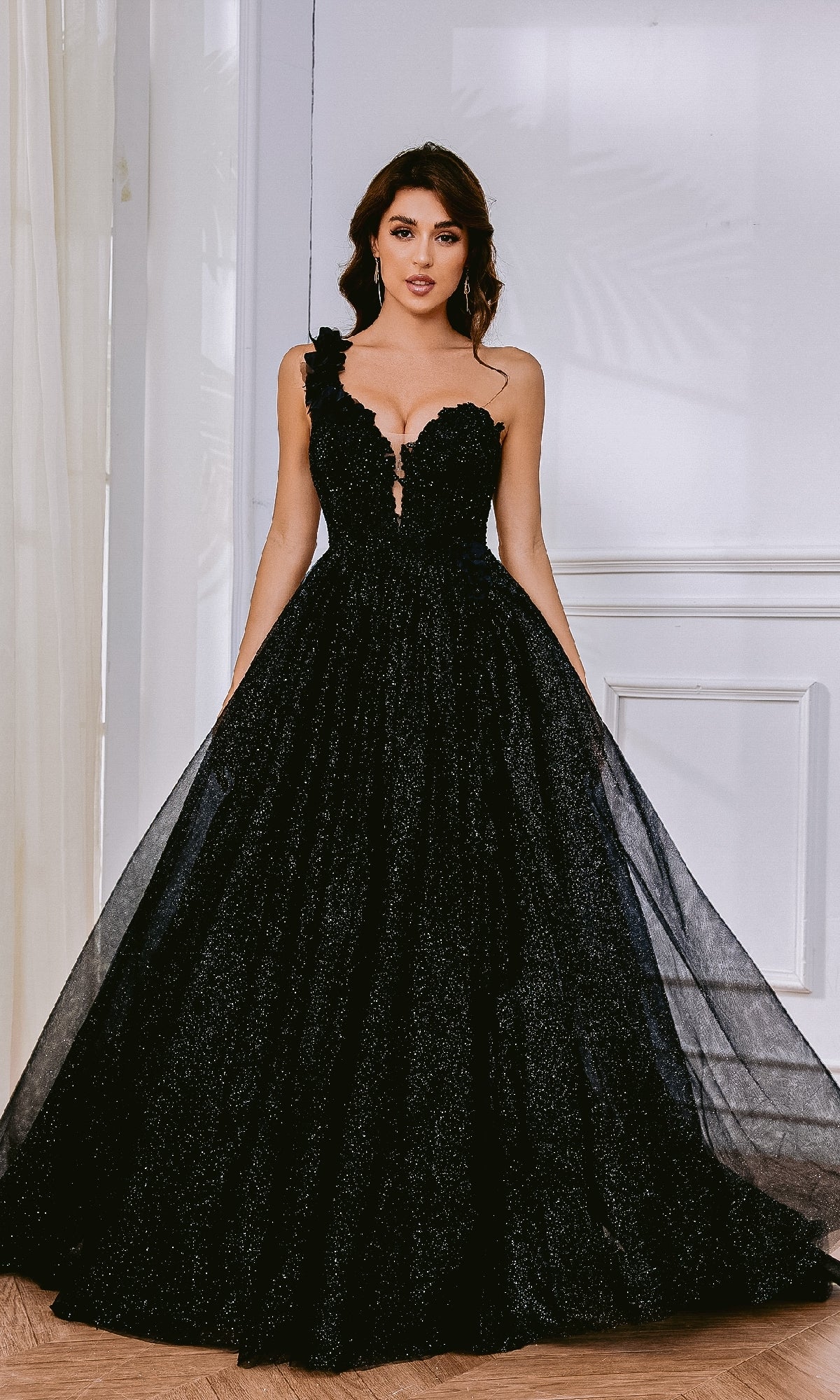 A woman in the Tina Holly TK554 One-Shoulder Glitter Prom Gown, featuring a black, floor-length design with a fitted bodice, full tulle skirt, and corset lace-up back, stands indoors against a white wall.