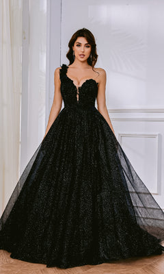 A woman in the Tina Holly TK554 One-Shoulder Glitter Prom Gown, featuring a black, floor-length design with a fitted bodice, full tulle skirt, and corset lace-up back, stands indoors against a white wall.
