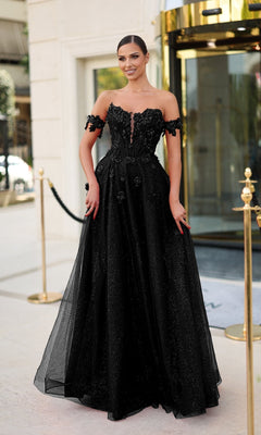 A woman wearing the Tina Holly TK582 Long Prom Dress, an elegant off-the-shoulder black gown with a corset bodice, stands outdoors before a building entrance with gold accents and glass doors.