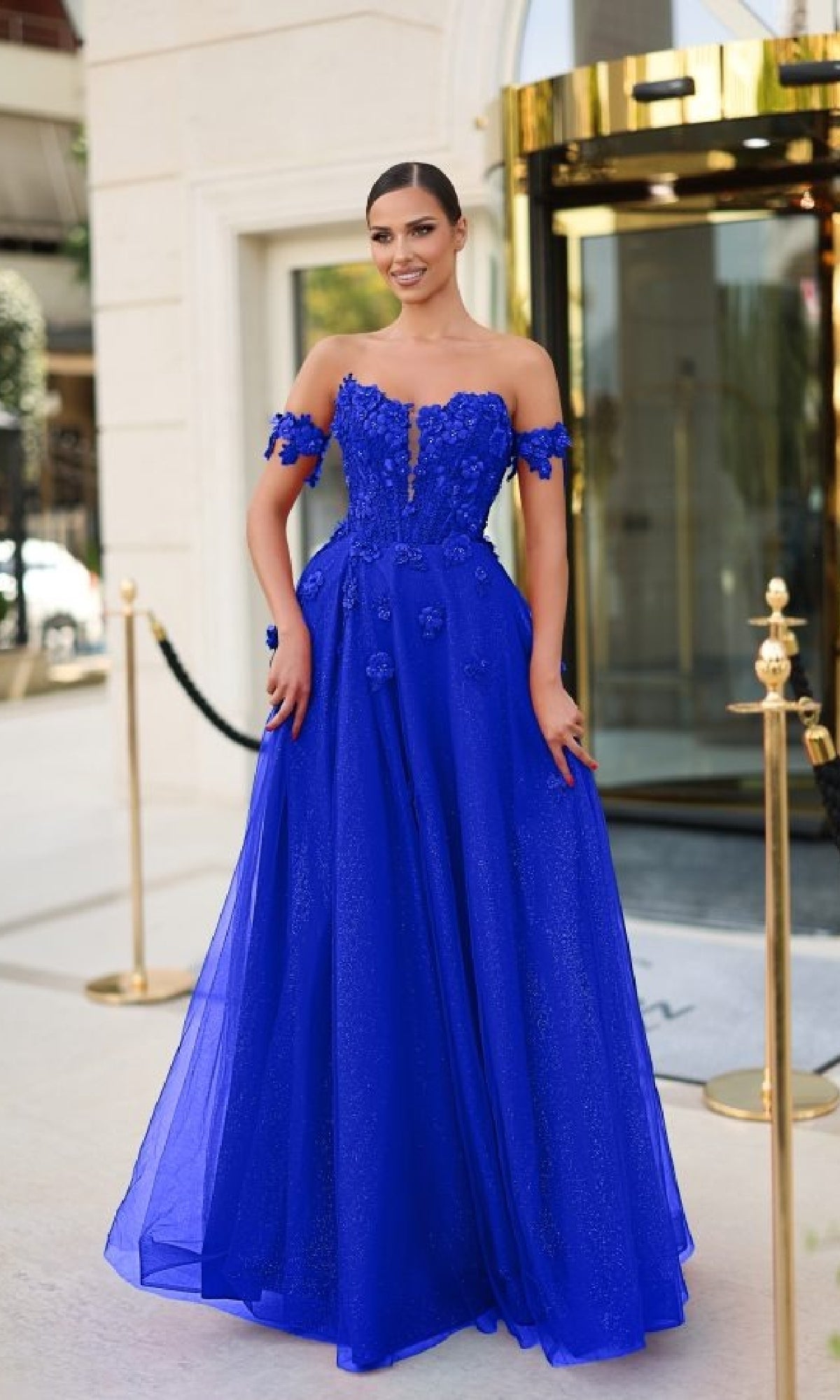 A woman wears the Tina Holly TK582, a royal blue, off-the-shoulder long prom dress with 3D flower appliques and a corset bodice, standing outside a building with glass doors and gold accents.