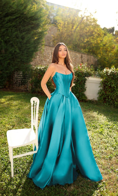 A woman in the Long Formal Dress: Tina Holly TK804, an Aqua Lake strapless mikado gown with a corset bodice, stands on grass beside a white chair amid greenery and sunlight.