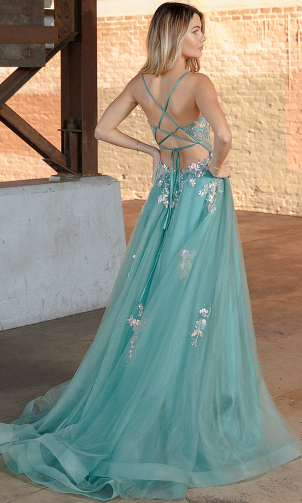 A woman in the Floral-Embellished Pastel Prom Ball Gown TM1003 poses gracefully in an industrial setting, showcasing its elegant strappy open back and delicate floral accents.