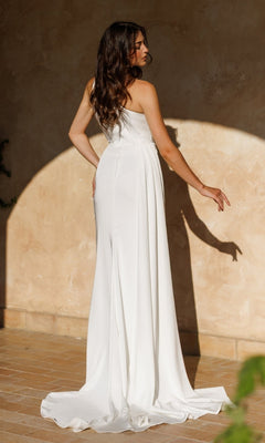 A woman with long brown hair wearing the One-Shoulder White Prom Gown: Tina Holly TT001W stands on a tiled floor, facing a textured beige wall as sunlight casts her shadow.
