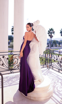 A woman models the Long Prom Dress: Tina Holly TT009B, a form-fitting gown with a thigh-high slit, standing beside a white angel statue on a balcony with ornate railings and palm trees in the background.