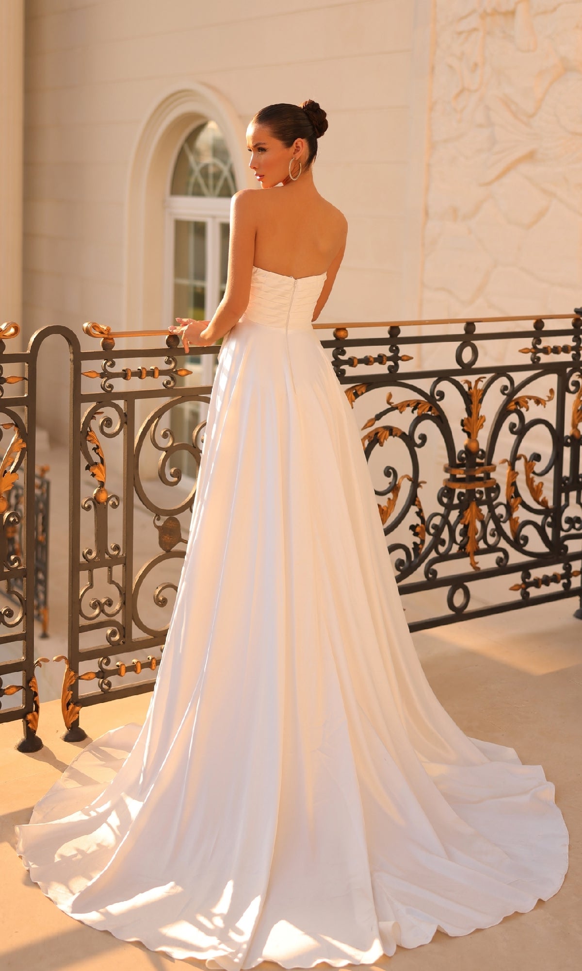 A woman wearing the Strapless Off-White Prom Gown: Tina Holly TT009 stands on a balcony with ornate black and gold railing, facing away from the camera.
