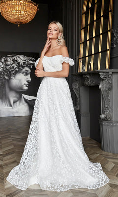 A woman wears the Long White Lace Formal Dress: Tina Holly TK023W, featuring floral patterns and an off-the-shoulder design, standing indoors near a fireplace with a statue and chandelier in the background.